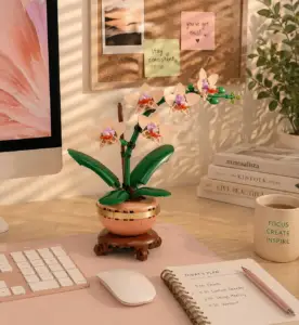 LEGO botanical orchid set on a pastel home office desk with pink desk mat, keyboard, notebook, coffee mug, and warm workspace decor.