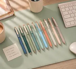 Pastel retractable pen set arranged on a green desk pad with gold accents, keyboard, coffee cup, sticky note, and modern office accessories.