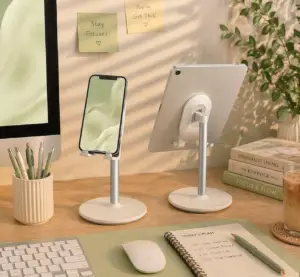 Adjustable white phone stand on a modern home office desk with tablet stand, keyboard, mouse, planner, iced coffee, and soft green workspace decor.